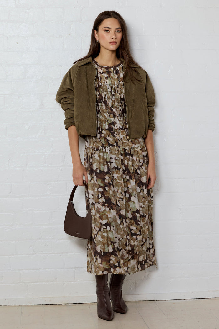 Woman wearing a floral dress, green jacket, and brown boots against a white brick wall.