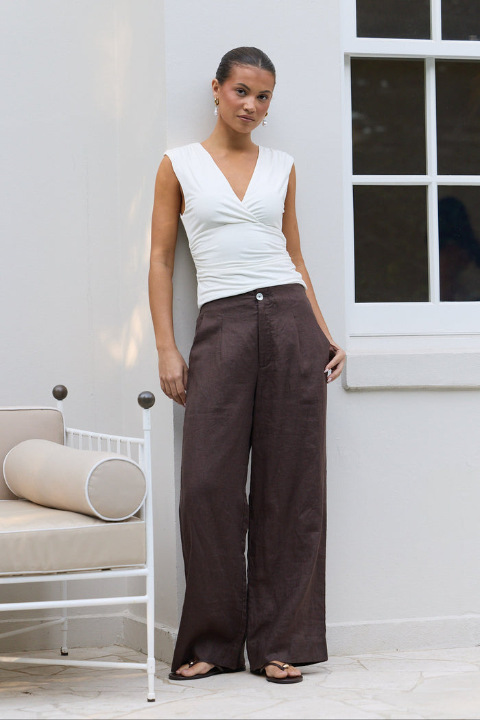 A woman stands outdoors against a white wall wearing a white sleeveless top and dark brown wide-leg trousers, with sandals on a patio next to a white chair.