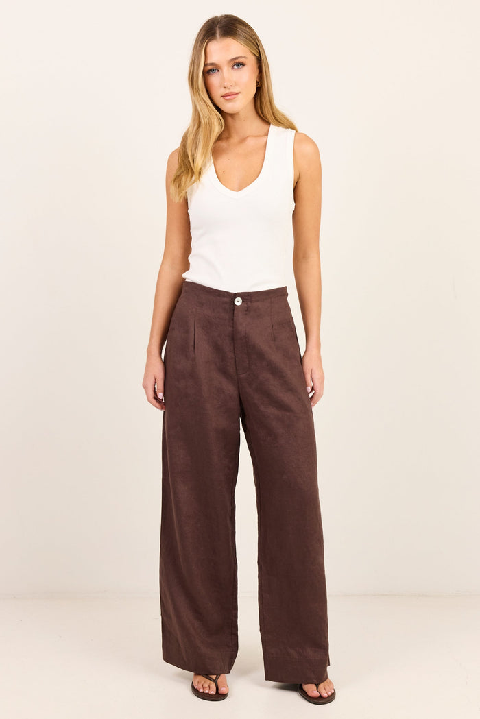 A woman modeling a pair of dark brown wide-leg trousers with a white sleeveless tank top, standing against a plain off-white studio background.
