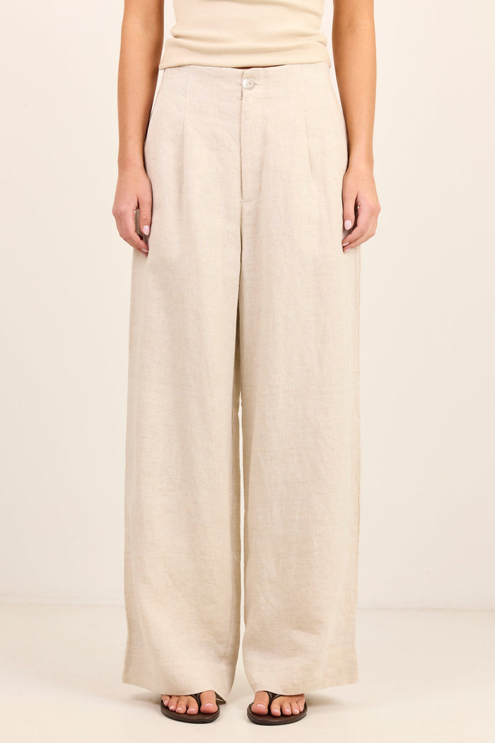A model wearing a pair of wide-leg linen pants in a light beige color, paired with a matching beige top and dark brown sandals, set against a plain cream background.