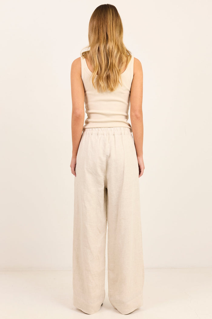 A woman with blonde hair is shown from the back, wearing a beige ribbed tank top and matching beige wide-leg linen pants against a plain off-white studio background.