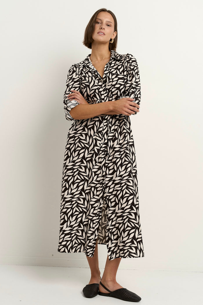 Woman wearing a black-and-white leaf-print midi dress with a relaxed button-down design and long sleeves, styled with black woven flats.
