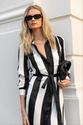 Woman wearing a black and white striped dress with sunglasses and clutch against a light-colored wall.