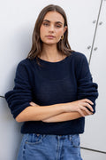 Woman wearing a navy sweater and blue jeans against a white wall.