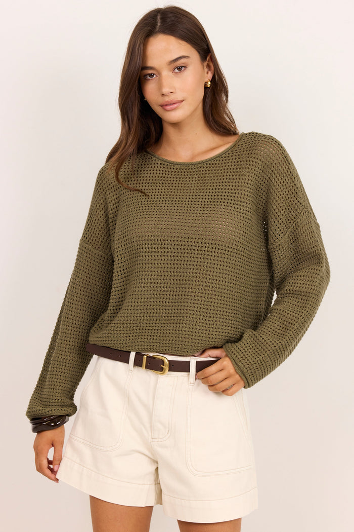 A woman modeling a textured olive green knit long-sleeve sweater paired with off-white shorts and a brown belt, set against a plain light background.