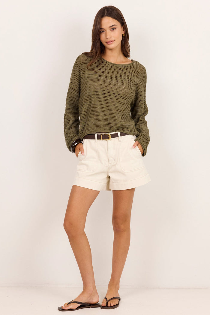 A woman modeling a long-sleeve olive green knit top, cream shorts, a thin brown belt, and black flip-flops against a plain off-white studio background.