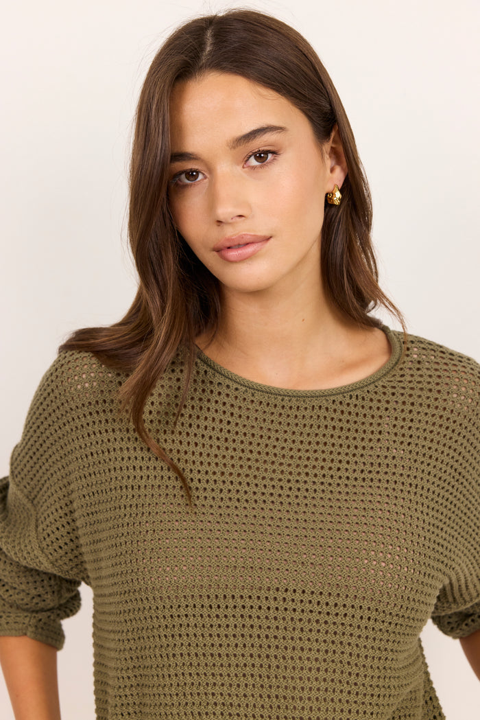A woman wearing a short-sleeve olive green open-knit crewneck top, photographed against a clean off-white background.