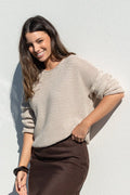 A woman posing outdoors against a white wall, wearing a beige knit sweater and a dark brown midi skirt, holding a small gray handbag.