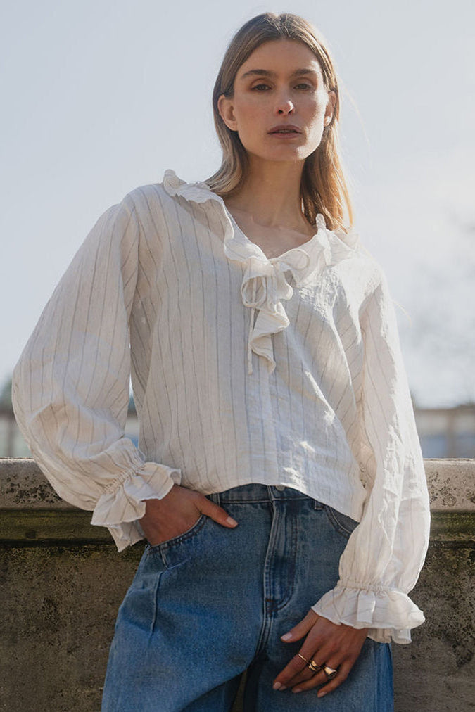Woman wearing a white blouse with ruffles and blue jeans standing against a cityscape background.