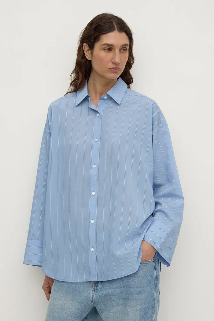 A woman wearing a light blue long-sleeve button-up shirt with subtle vertical stripes, paired with light-wash blue jeans, standing against a plain off-white background.