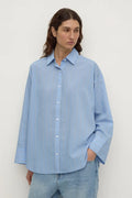 A woman wearing a light blue long-sleeve button-up shirt with subtle vertical stripes, paired with light-wash blue jeans, standing against a plain off-white background.