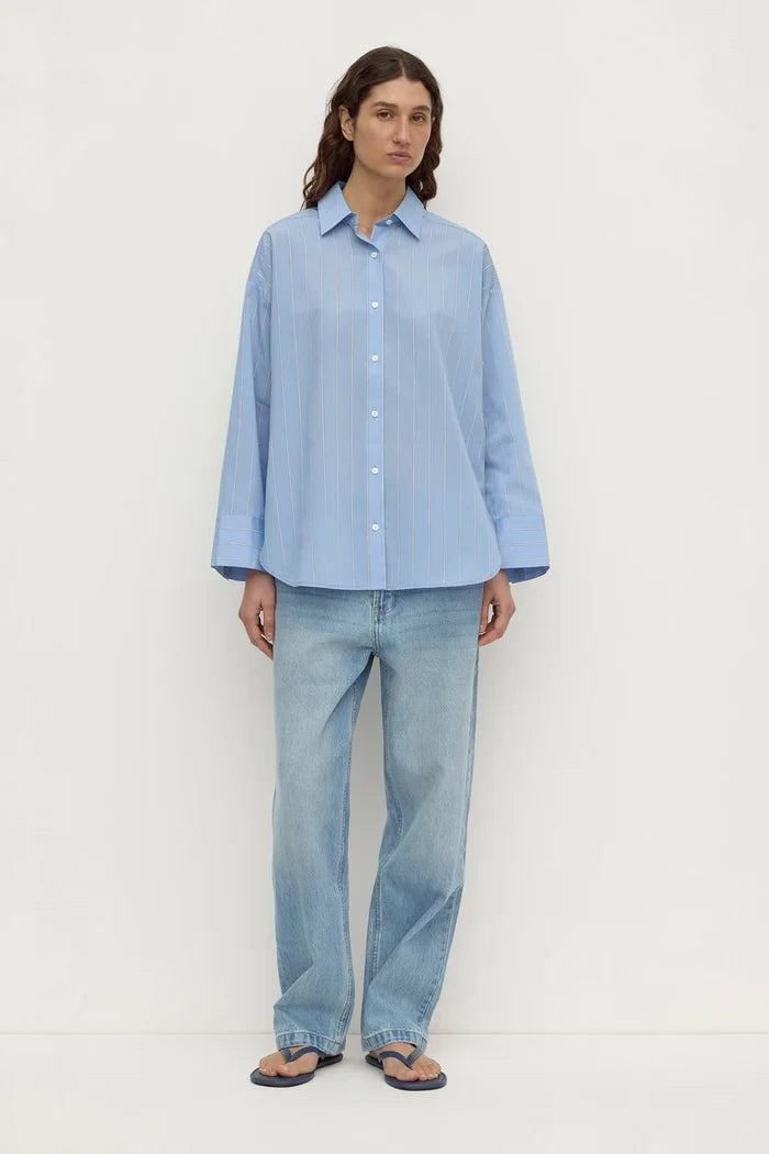 A woman stands against a plain off-white studio background wearing a light blue button-up shirt with subtle vertical stripes, light-wash blue jeans, and dark flip-flop sandals.