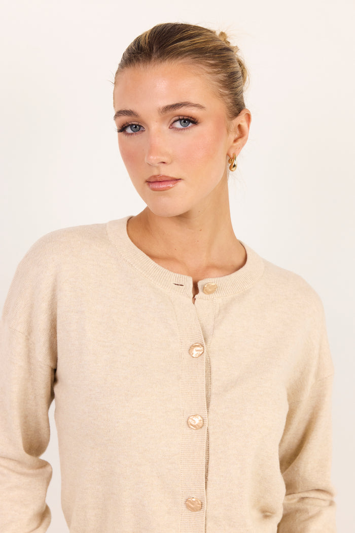 A woman wearing a light beige knit cardigan sweater with gold buttons, photographed against a plain off-white studio background.