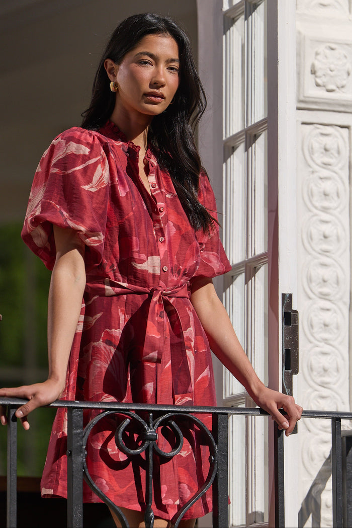 A woman stands on a balcony wearing a red patterned short-sleeve dress with a tie waist. The dress has a pinkish-red base with lighter abstract floral or marbled patterns.