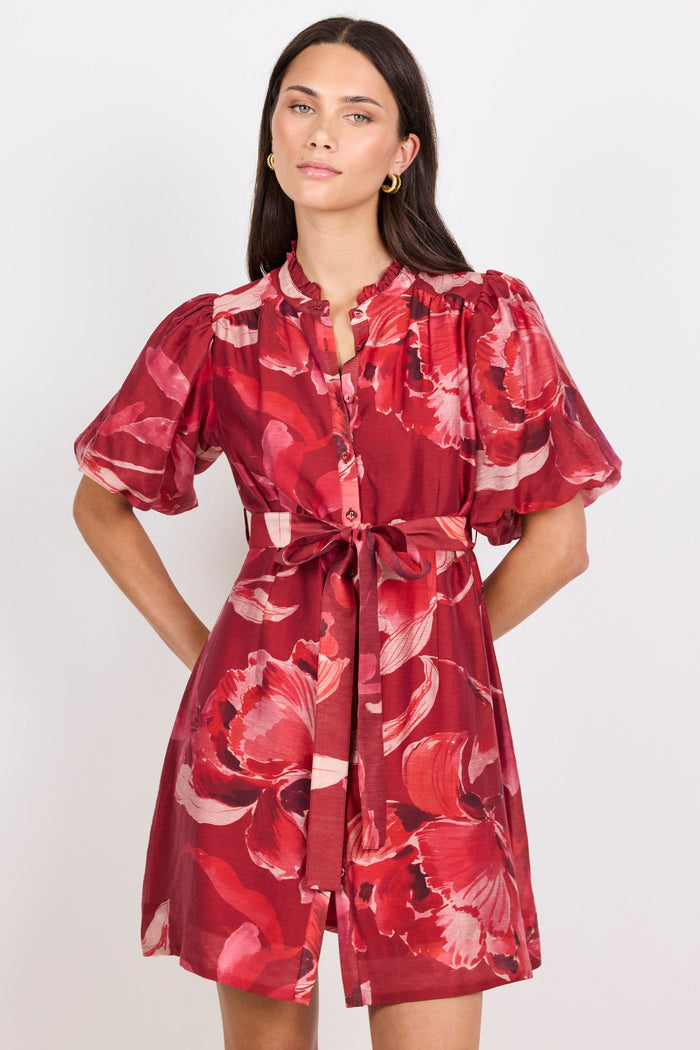 A woman modeling a red floral print mini dress with puff sleeves, a button-up front, and a matching waist tie, set against a plain light gray background.