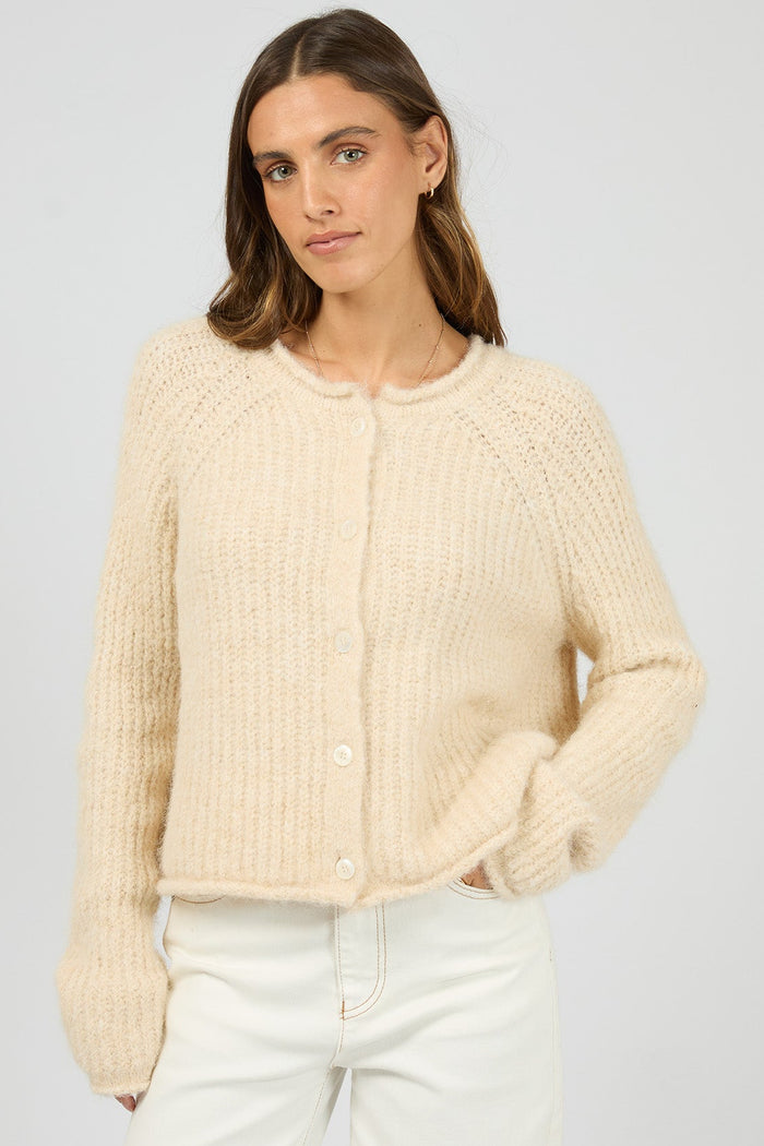Woman wearing a beige knitted cardigan and white pants on a light gray background