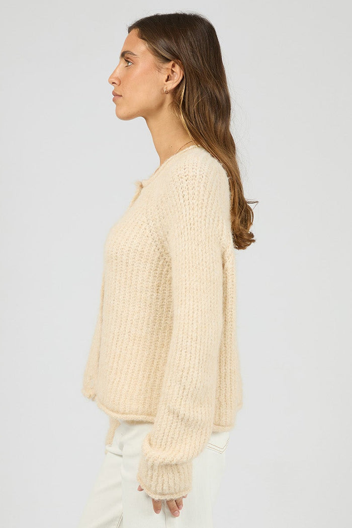 Woman wearing a beige ribbed sweater and white pants on a plain background