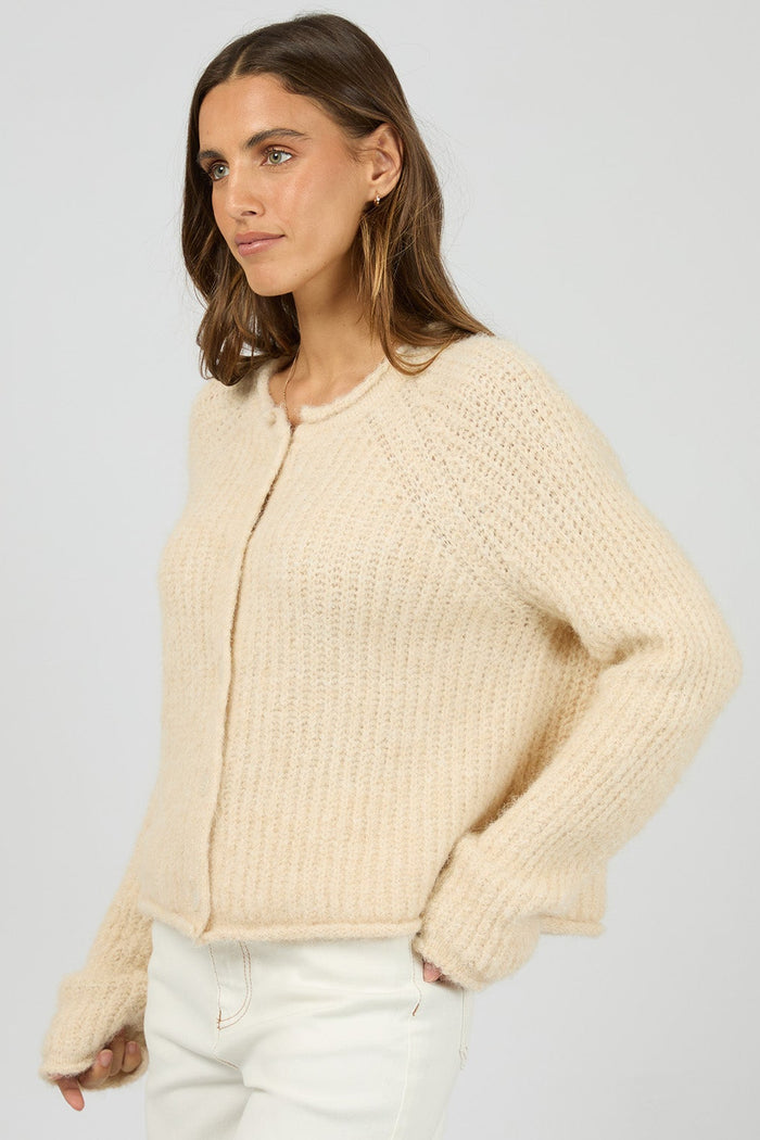 Woman wearing a beige cardigan and white pants on a light gray background