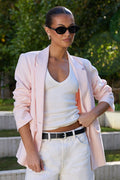 A woman wearing a light pink blazer, white tank top, and white wide-leg pants with a black belt, standing outdoors in front of green trees and concrete steps.
