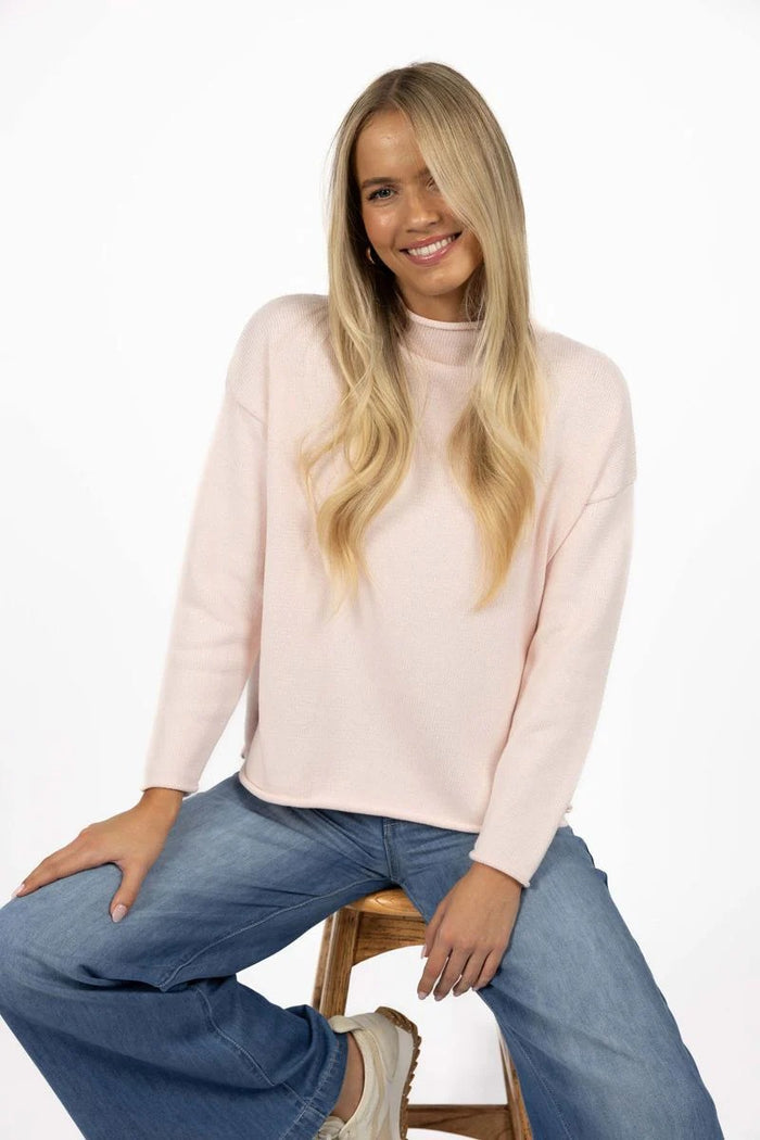 Woman wearing a light pink sweater and blue jeans sitting on a wooden stool against a white background
