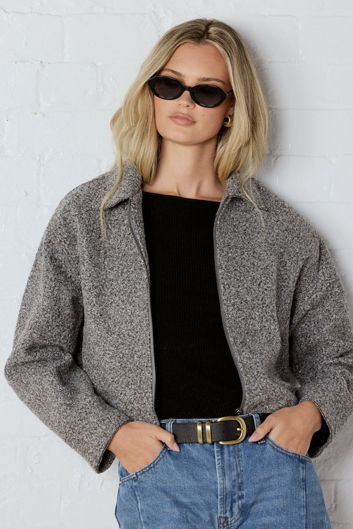 Woman wearing a gray jacket, black top, and blue jeans against a white wall.
