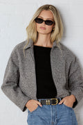 Woman wearing a gray jacket, black top, and blue jeans against a white wall.