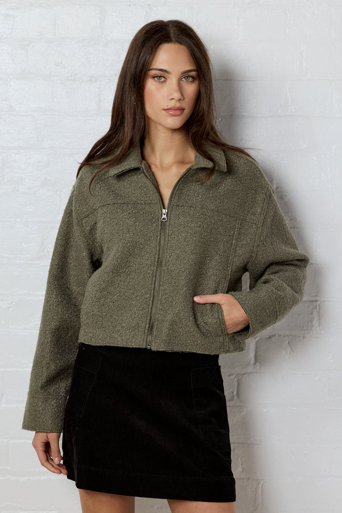 Woman wearing a green jacket and black skirt against a white brick wall.