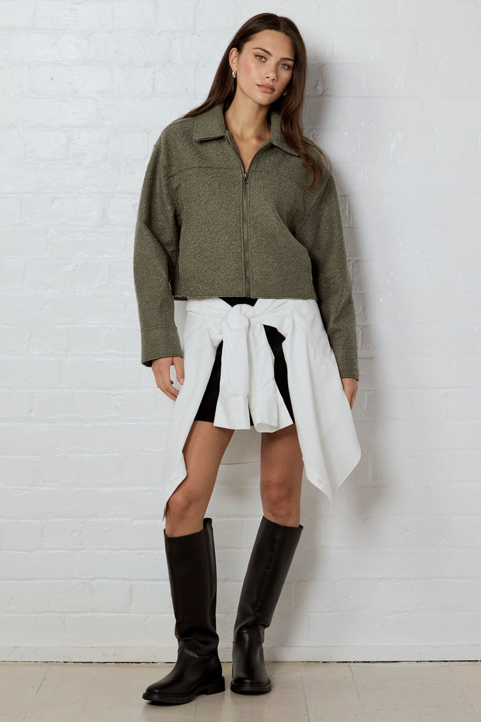 A woman stands against a white brick wall wearing an olive green cropped button-up shirt, a white shirt tied around her waist, black shorts, and black knee-high boots.