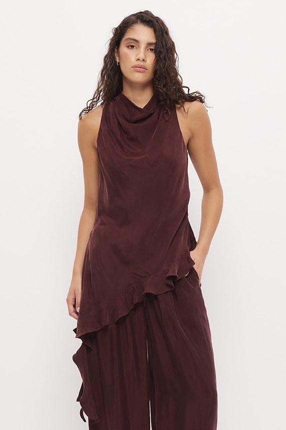 A woman modeling a sleeveless, dark burgundy draped top and matching wide-leg pants set, paired with black flip-flops, against a plain white studio background.