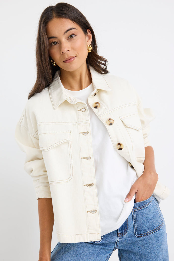 A woman modeling a cream-colored denim jacket with gold-tone buttons over a white t-shirt and blue jeans, set against a plain white studio background.