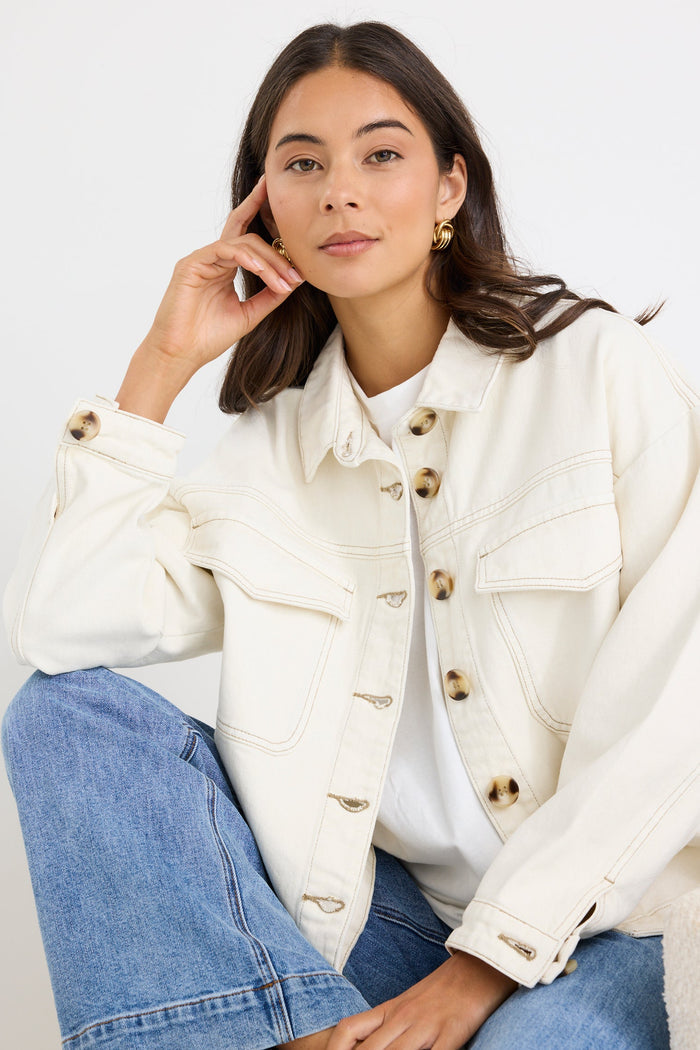 A woman wearing a cream-colored denim jacket with gold-tone buttons and blue jeans, posing against a clean white studio background.