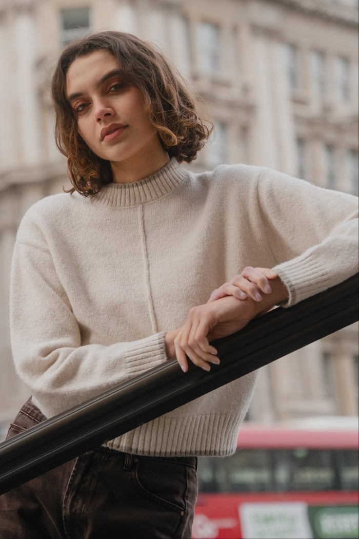 Person wearing a beige sweater standing in front of a building with a blurred red bus in the background