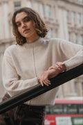 Person wearing a beige sweater standing in front of a building with a blurred red bus in the background