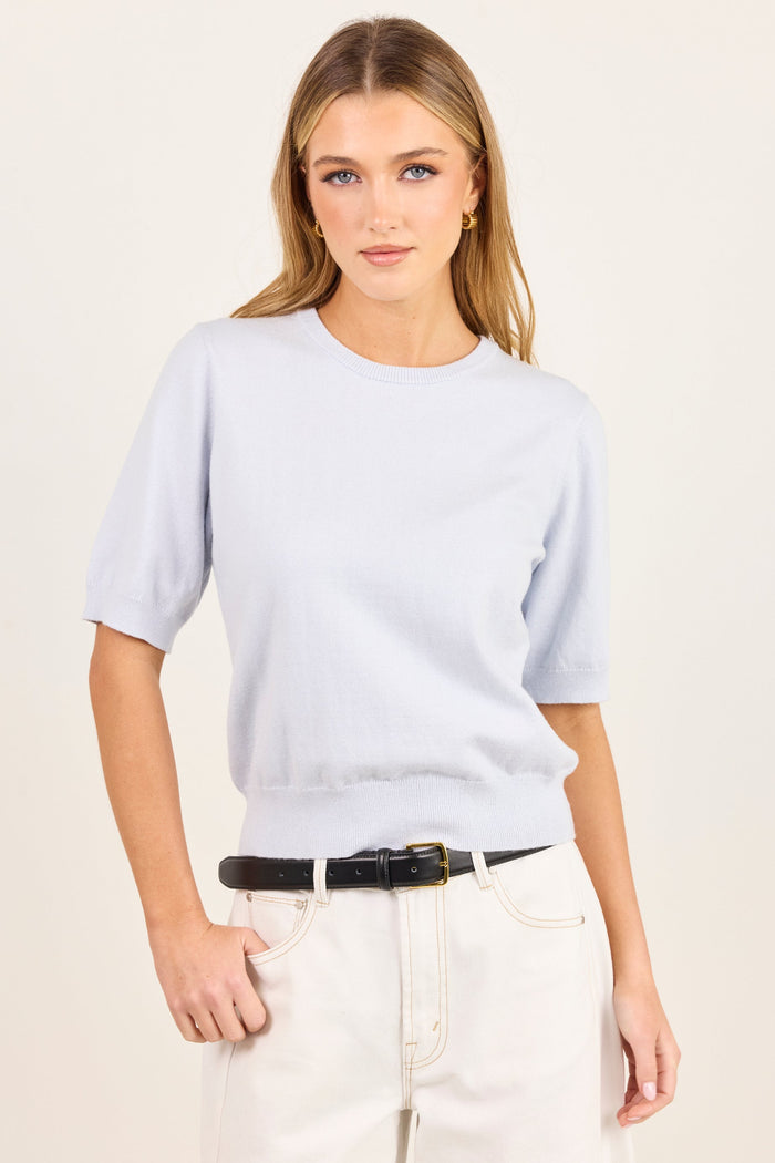 A woman modeling a light blue short-sleeve crewneck knit top, paired with cream-colored pants and a black belt, against a plain off-white studio background.