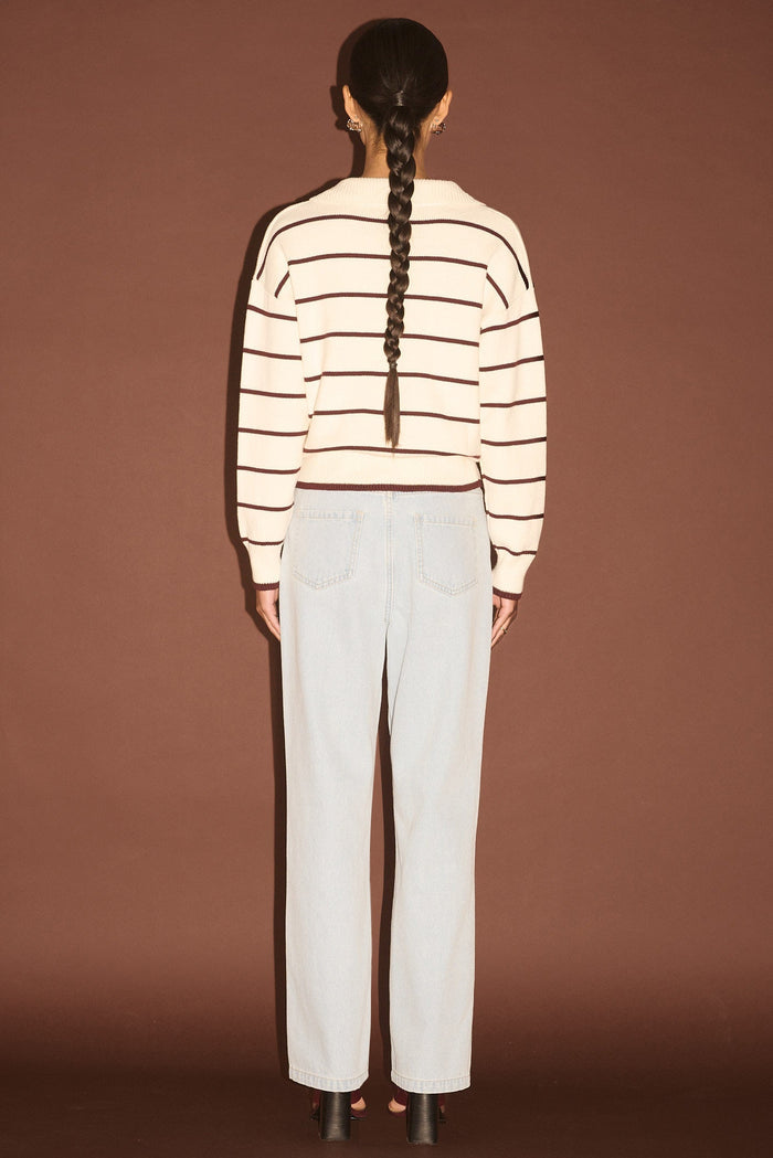 Person wearing a striped sweater and white pants against a brown background