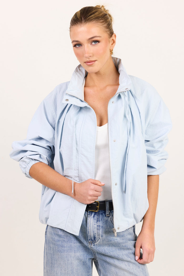 A woman modeling a light blue oversized button-up shirt jacket over a white tank top and light-wash jeans, photographed against a plain off-white studio background.