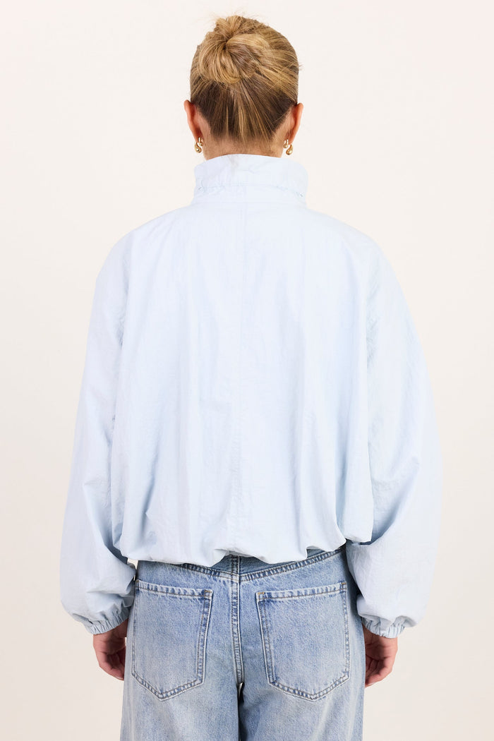A person viewed from the back wearing a light blue high-neck long-sleeve shirt and light-wash blue jeans against a plain off-white studio background.