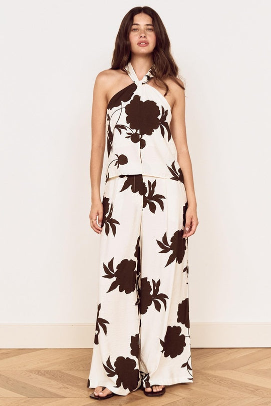 A woman modeling a halter-neck two-piece set with wide-leg pants, featuring a cream base with a large dark brown floral print, set against a plain off-white wall and light wood flooring.