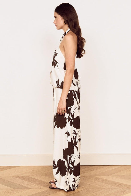 A woman stands in profile wearing a sleeveless halter maxi dress with a bold dark brown floral print on an off-white background, set against a plain white wall and light wood herringbone floor.
