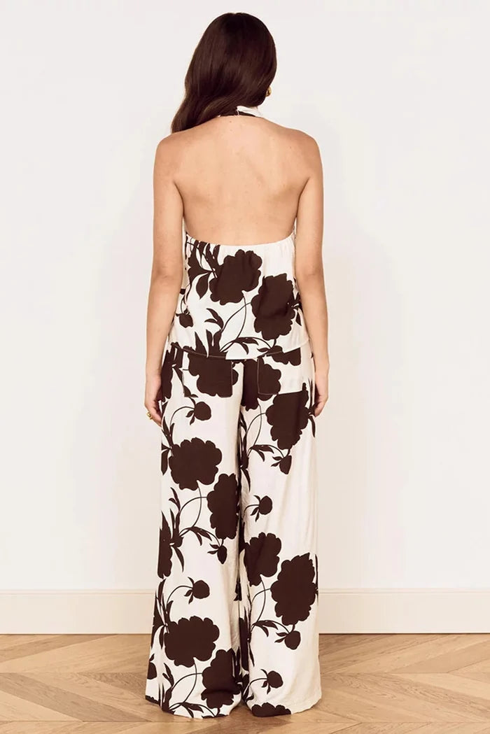 A woman viewed from behind wearing a strapless halter-style jumpsuit with a bold dark brown floral print on an off-white background, standing against a plain white wall on a light wood floor.