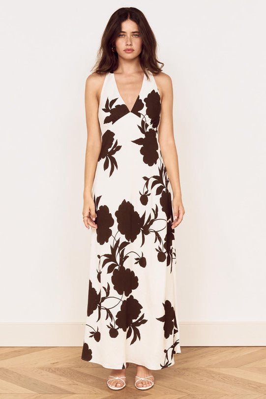 A woman modeling a sleeveless halter-neck maxi dress with a dark brown floral print on an off-white background, standing against a plain white wall on a light wood floor.