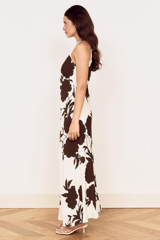 A woman stands in profile wearing a sleeveless maxi dress with a bold dark brown floral print on an off-white background, paired with clear high-heeled sandals, set against a plain white wall and light wood parquet floor.