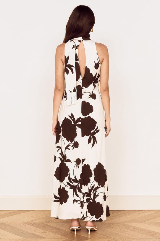 A woman viewed from the back wearing a sleeveless halter maxi dress with a large dark brown floral print on an off-white background, standing in a room with a light wall and wood floor.