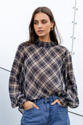 Woman wearing a plaid shirt and blue jeans standing against a white wall.