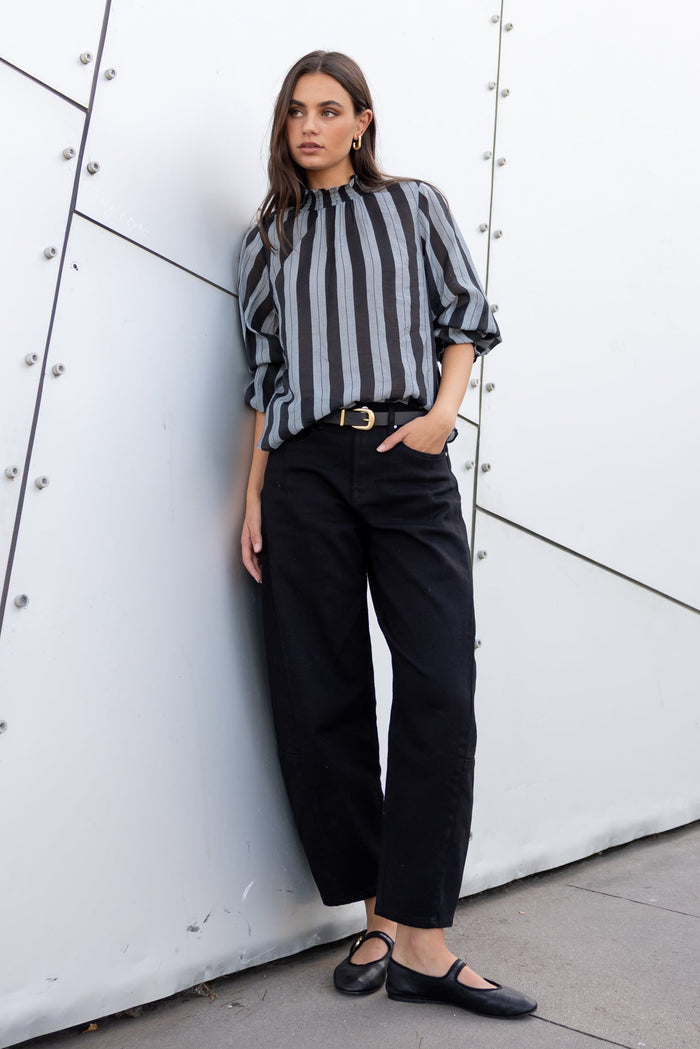 Woman in a striped shirt and black pants leaning against a modern architectural wall.