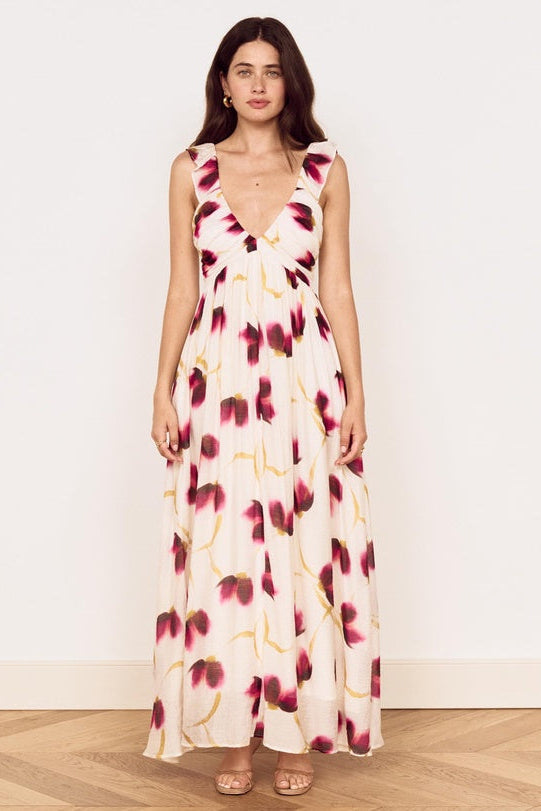 A woman modeling a sleeveless, floor-length cream maxi dress with a dark red and gold floral print, standing against a plain white wall on a light wood floor.