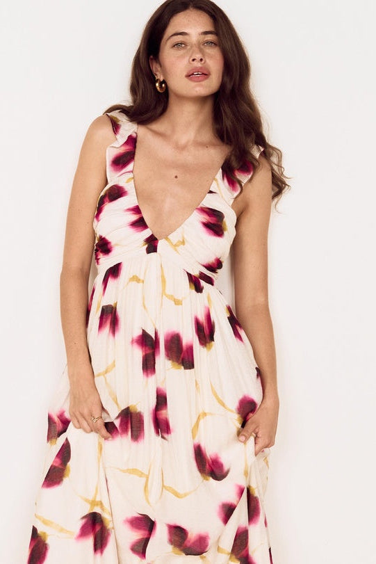 A woman modeling a sleeveless, deep V-neck maxi dress with a cream base and a dark pink and yellow abstract floral pattern, set against a plain white background.