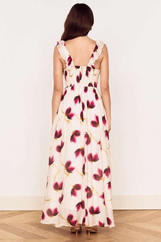 A woman viewed from behind wearing a sleeveless maxi dress with a cream base and a dark red and gold floral pattern, standing on a light wood floor against a white wall.