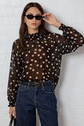 Woman wearing a sheer polka dot blouse and jeans against a white brick wall.