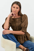 Woman sitting on a white chair wearing a brown checkered blouse and blue jeans.